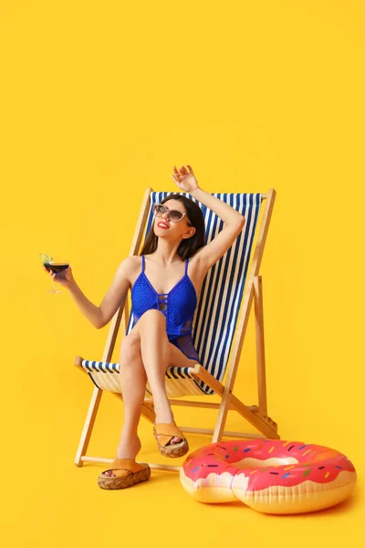 Beautiful young woman with cocktail sitting on deck chair against color background