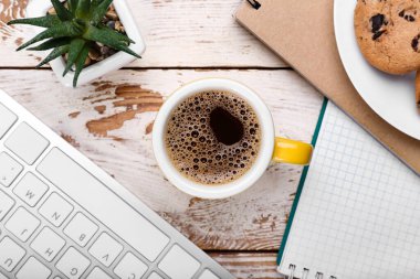 Bir fincan kahve, klavye, defterler ve hafif ahşap arka planda saksı.