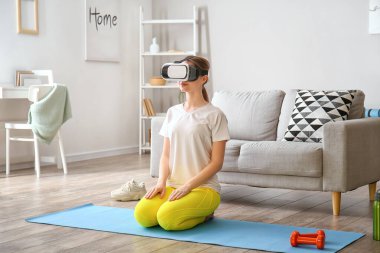 Sanal gerçeklik gözlüklü genç kadın evde yoga yapıyor.
