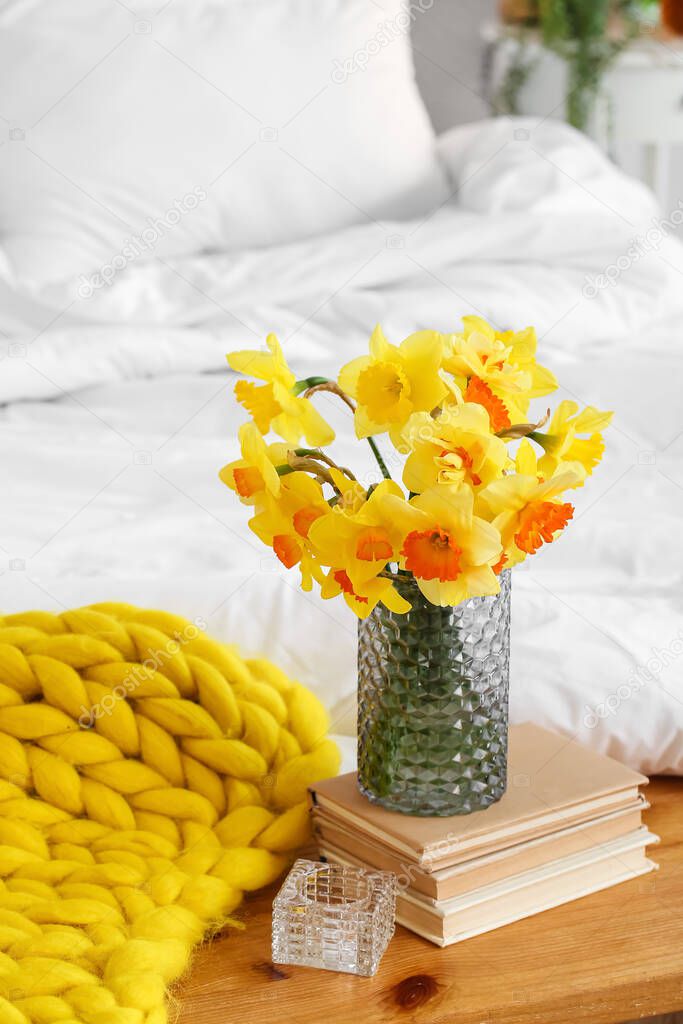 Vase with narcissus flowers and books on bedside bench in room
