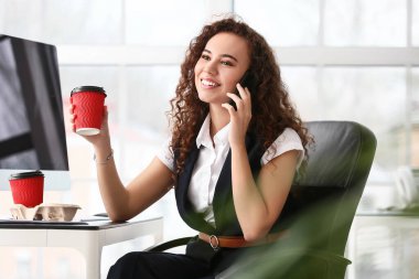 Ofiste elinde kahve olan genç bir kadın cep telefonuyla konuşuyor.
