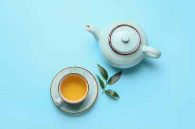 Teapot and cup of tea with green leaves on color background