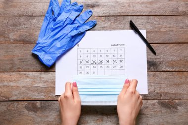 Tıbbi maskeli kadın eller, takvim, lastik eldivenler ve tahta arka planda kalem.