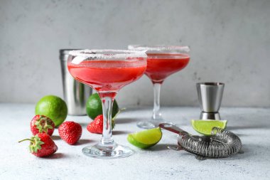 Glasses of strawberry daiquiri cocktail, limes, berries and strainer on light background