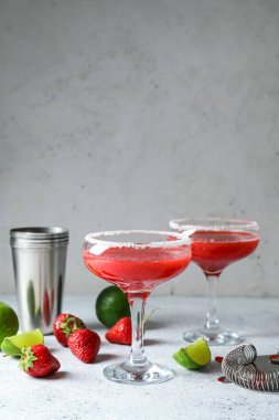 Glasses of strawberry daiquiri cocktail, limes, berries, shaker and strainer on light background