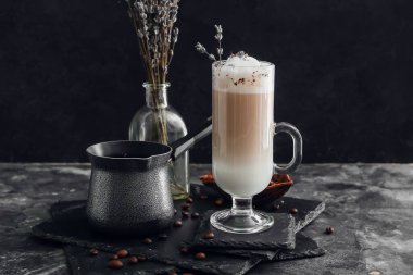 Bir bardak lavantalı latte, kahve demliği ve koyu arkaplanda çiçekler.