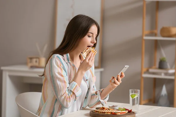 Mooie Jonge Vrouw Met Mobiele Telefoon Eten Smakelijke Quesadilla Thuis — Stockfoto