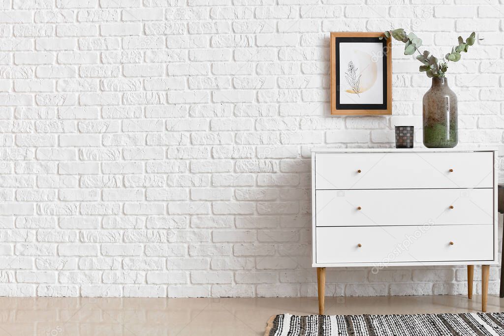 Modern chest of drawers with vase and candle near brick wall