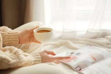 Evde cep telefonu kullanan bir fincan kahveli genç kadın, yakın plan.