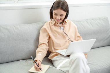 Dizüstü bilgisayarı olan genç bir kadın evde deftere bir şeyler yazıyor.