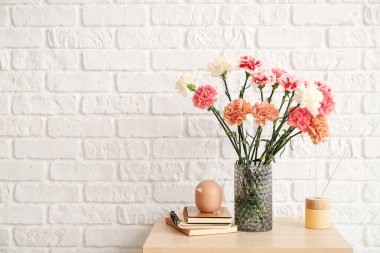 İçinde karanfil çiçekleri, kitaplar ve sazlıklar olan vazo tuğla duvarın yanında.