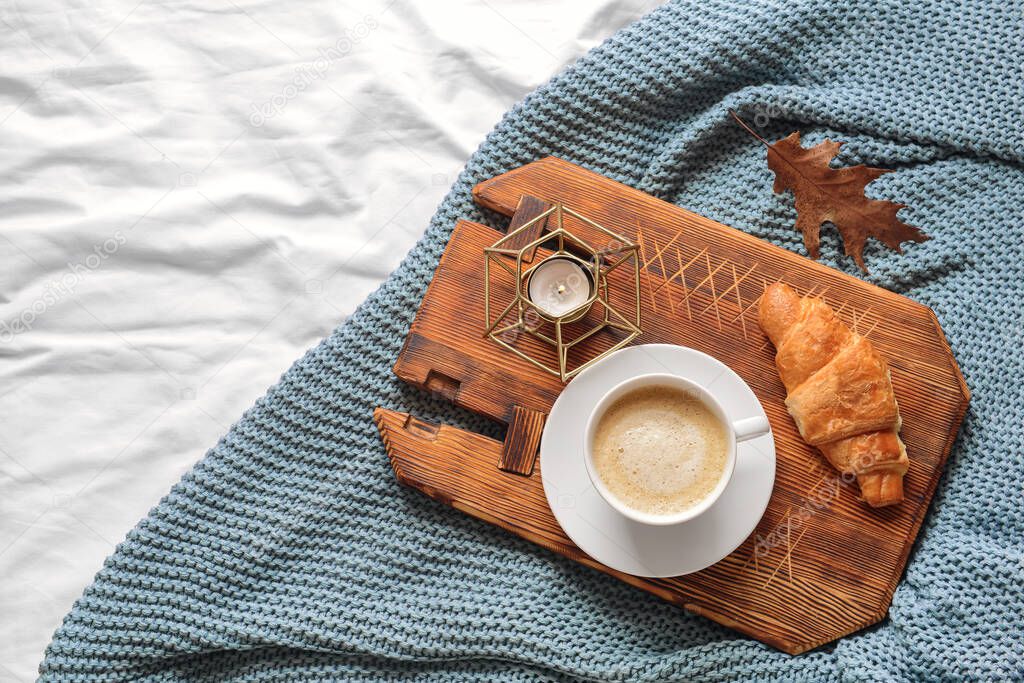 Board with cup of coffee, croissant and candle on knitted plaid