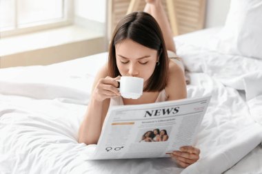 Gazeteli genç bir kadın yatak odasında kahve içiyor.