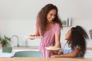 Annesi ile birlikte mutfakta taze salata yiyen Afro-Amerikalı küçük kız.