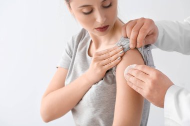 Doktor, arka planda genç bir kadının koluna pamuk pedi takıyor.