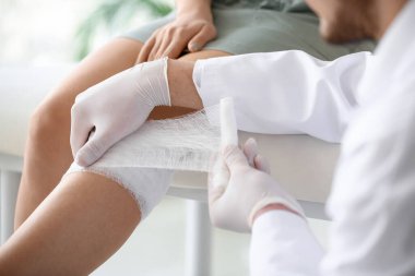 Doktor klinikteki genç bayanın dizine bandaj yapıştırıyor.