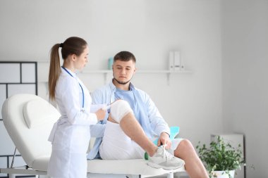 Doktor, klinikteki genç adamın dizine bandaj yapıştırıyor.
