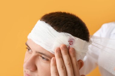 Doktor, arka planda genç bir adamın kafasına bandaj yapıştırıyor.