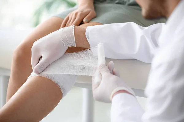 Doctor applying bandage onto knee of young woman in clinic