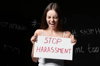 Elinde kağıt olan stresli genç bir kadın koyu arkaplanda DUR HARASSMENT