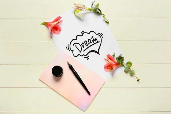 Paper with word DREAM, flowers, notebook and ink on white wooden background