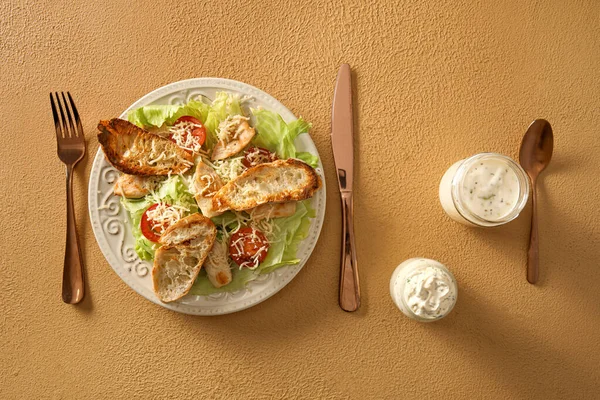 Plate of tasty Caesar salad and jars with sauce on beige background