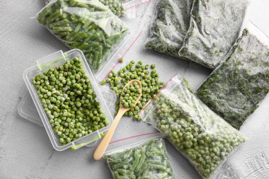 Gri arka planda farklı donmuş sebzelerle plastik kap ve çanta.