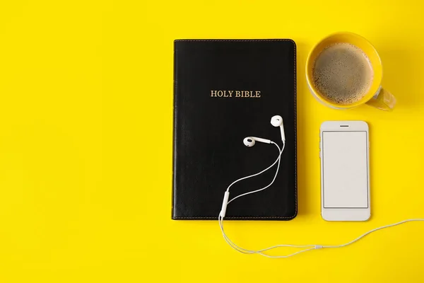 Holy Bible, earphones, cup of coffee and mobile phone on color background
