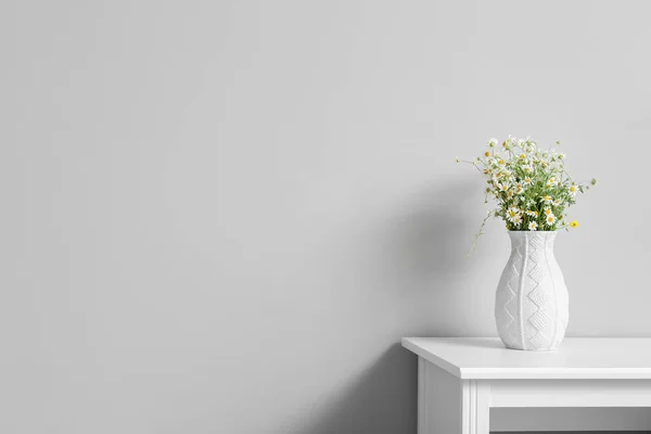 Vase with chamomiles on table near light wall