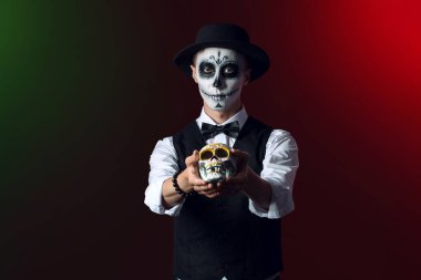 Meksika Ölüler Günü (El Dia de Muertos) için koyu arkaplanda boyalı yüzü ve şeker kafatası olan genç bir adam.