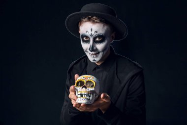 Meksika Ölüler Günü (El Dia de Muertos) için koyu arkaplanda boyalı yüzü ve şeker kafatası olan genç bir adam.