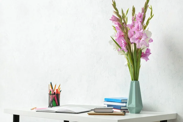 Vase with beautiful gladiolus flowers, books and stationery on table against light wall