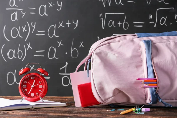 School backpack with stationery and alarm clock on table in classroom