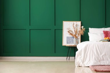 Renk duvarının yanındaki masada çiçekler olan rahat bir yatak ve vazo.