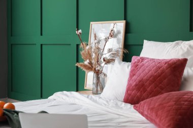 Yumuşak yastıklı yatak, vazodaki çiçekler renk duvarının yanındaki masada, yakın plan.