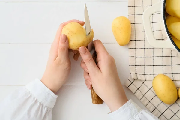Woman peeling raw potatoes on light wooden background, closeup