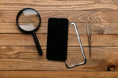 Tahta arka planda parçalanmış cep telefonu, büyüteç ve cımbız.