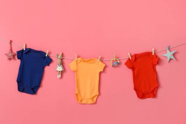 Farklı bebek kıyafetleri ve pembe arka planda asılı duran oyuncaklar.