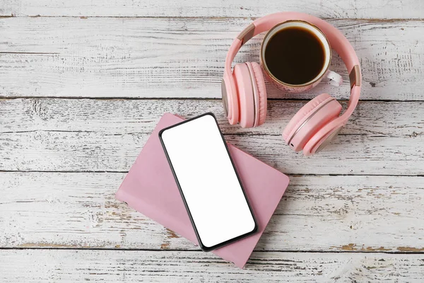 Modern mobile phone, headphones, notebook and cup of coffee on light wooden background