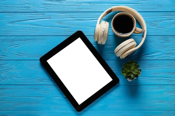 Modern tablet computer, headphones, cup of coffee and houseplant on color wooden background