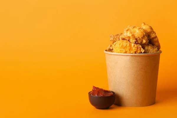 Paper bucket with tasty popcorn chicken and tomato sauce on color background