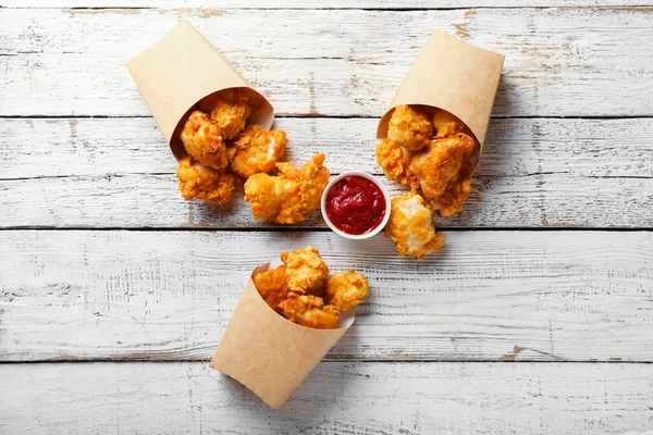 Paper boxes with tasty popcorn chicken and tomato sauce on light wooden background