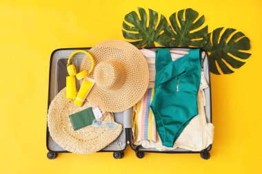 Sarı arka planda giysiler, mayolar ve plaj aksesuarları olan bavulu aç, kapat.