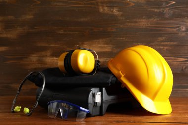 Sert şapka, güvenlik gözlükleri, koruyucu kulaklıklar ve tahta arka planda alet çantası.