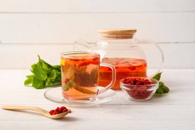 Bir bardak sıcak goji çayı ve beyaz ahşap arka planda nane ve böğürtlen.