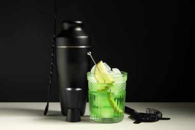 Siyah duvarın yanındaki beyaz masada bir bardak taze kokteyl ve barmen aletleri.