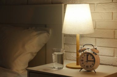 Çalar saat, parlayan lamba ve karanlık yatak odasındaki masada bir bardak su.