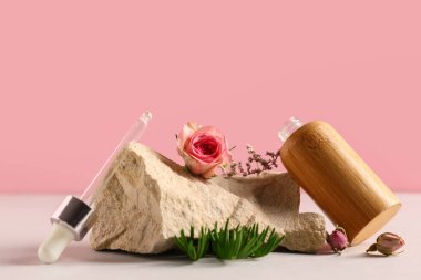 Temel yağ şişesi, dekoratif taş ve pembe duvarın yanındaki beyaz masanın üzerindeki çiçeklerle kompozisyon, yakın plan.