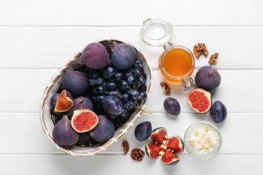 Hasır sepetli kompozisyon, olgun incirler, beyaz ahşap arka planda üzüm ve bal kavanozu.