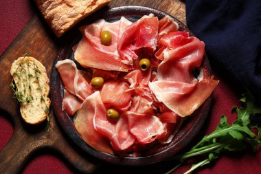 Lezzetli prosciutto dilimleri, zeytin ve kırmızı arkaplanda roka yaprakları.